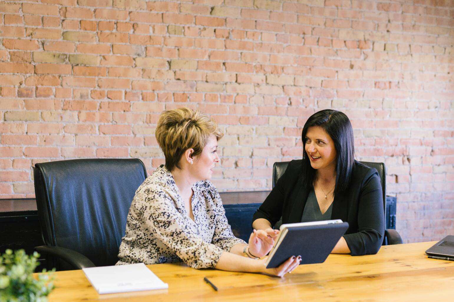 Two businesswomen are sitting at a conference table having a discussion about taxes while looking at a tablet.