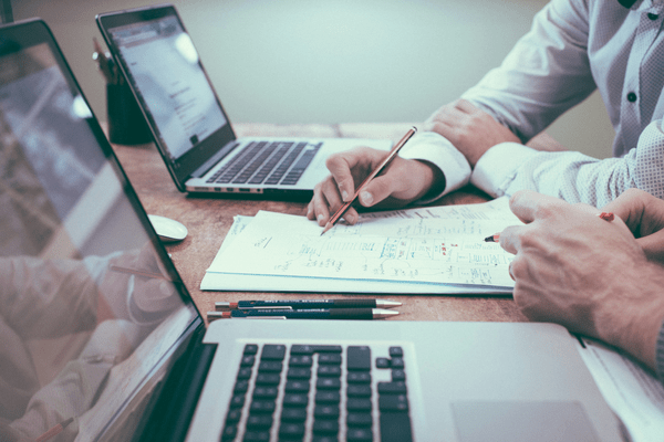 A person is seen using a pencil to make notes on a document while another person looks on. Two open laptops are on the table. The professional is working with a financial planning client.